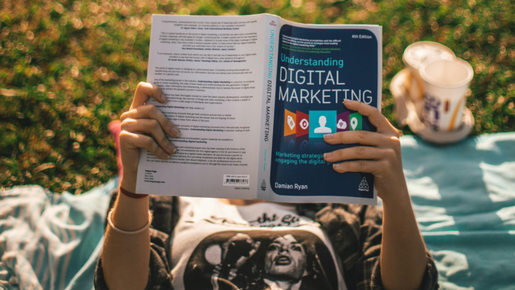 A person lies on a blanket in the grass, reading "Understanding Digital Marketing." A drink in a cup is beside them.