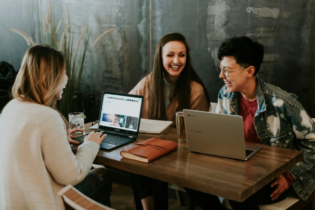 A group of people smiling and sitting around a table working on laptops.