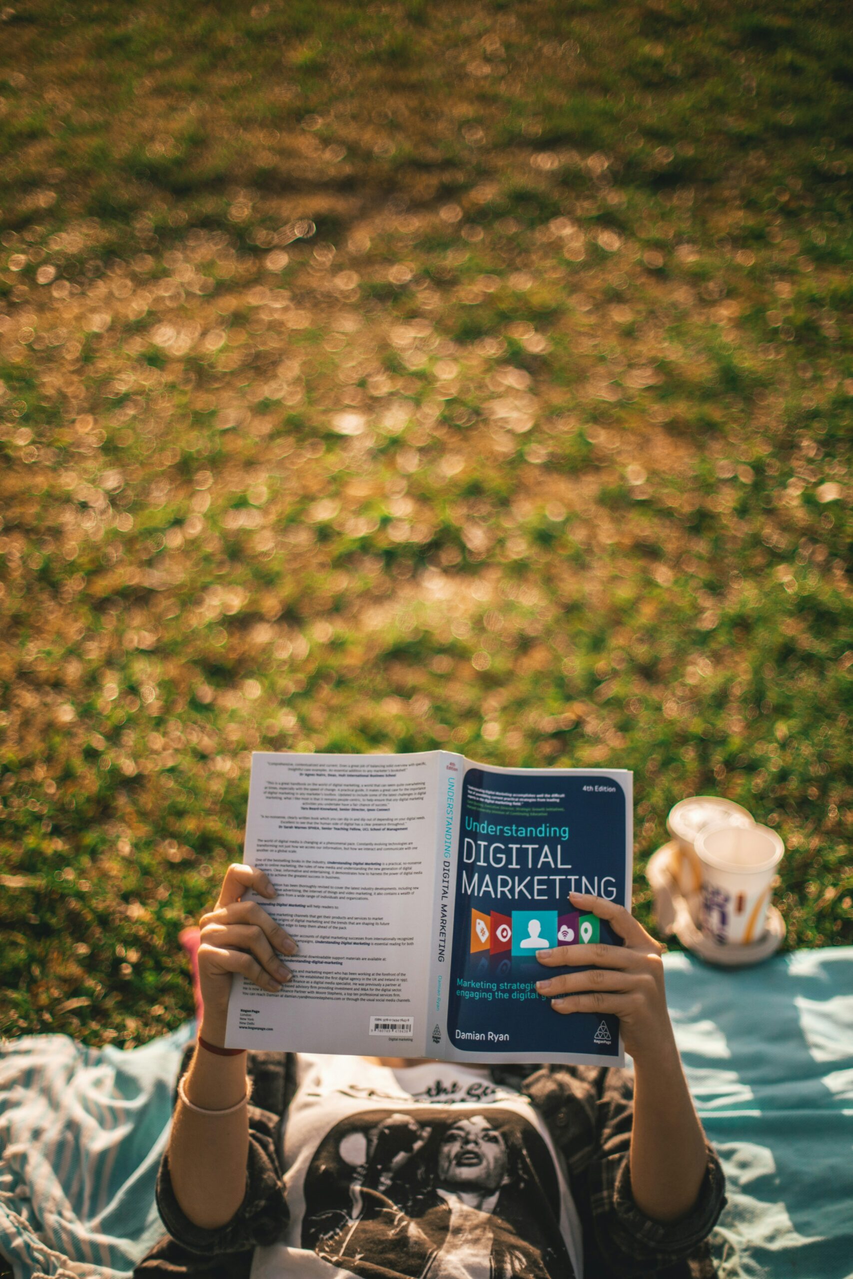 A person lies on a blanket in the grass, reading "Understanding Digital Marketing." A drink in a cup is beside them.