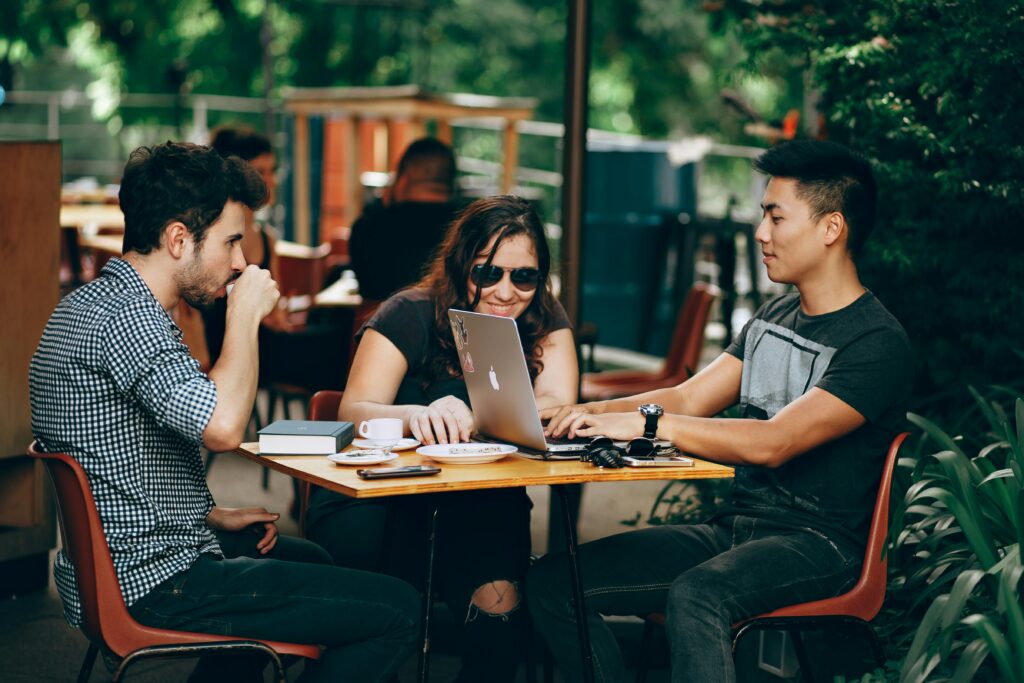 a group of people sat around a table at a coffee shop, working together on a laptop discussing Outsourcing press release distribution.