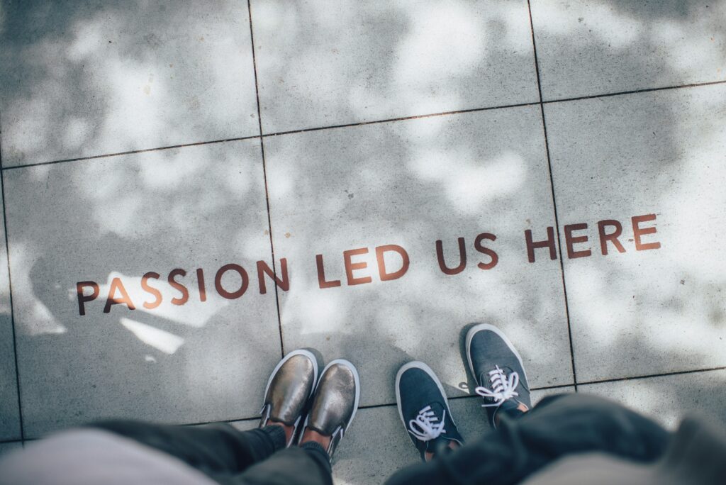 An image showing a pavement with 'Passion led us here' written on it. Two pairs of legs can be seen standing next to the text