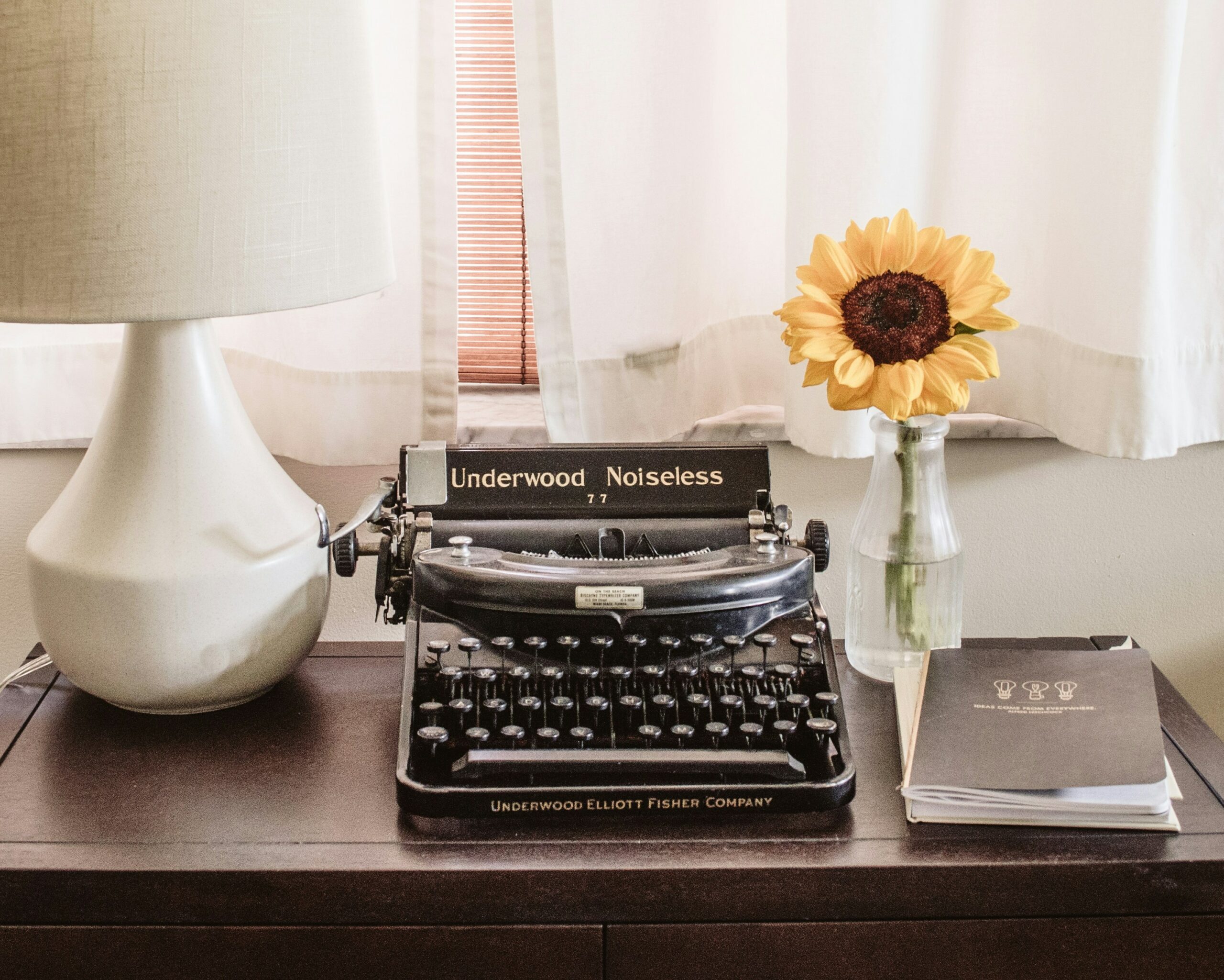 A photo of a type writer on a desk with a flower and lamp beside it.